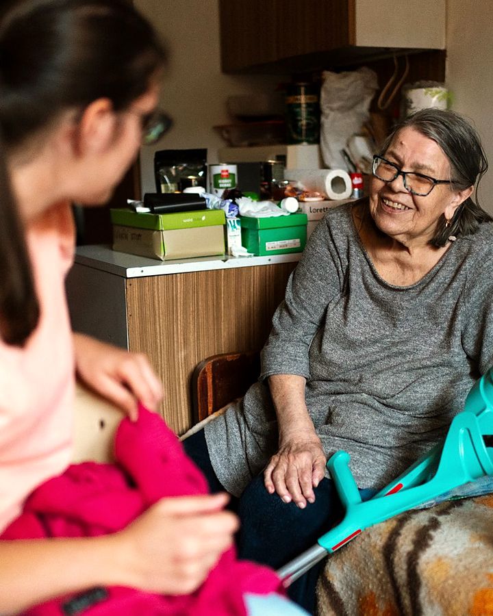 Una cuidadora acompaña de cerca a una señora mayor, sentadas en casa