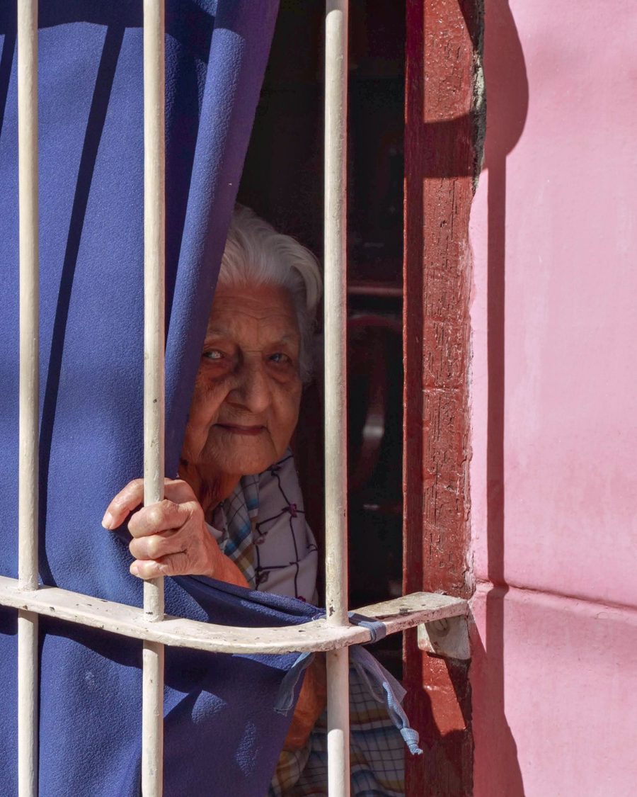 Una mujer mayor asomada a la ventana del cuarto, entre las cortinas